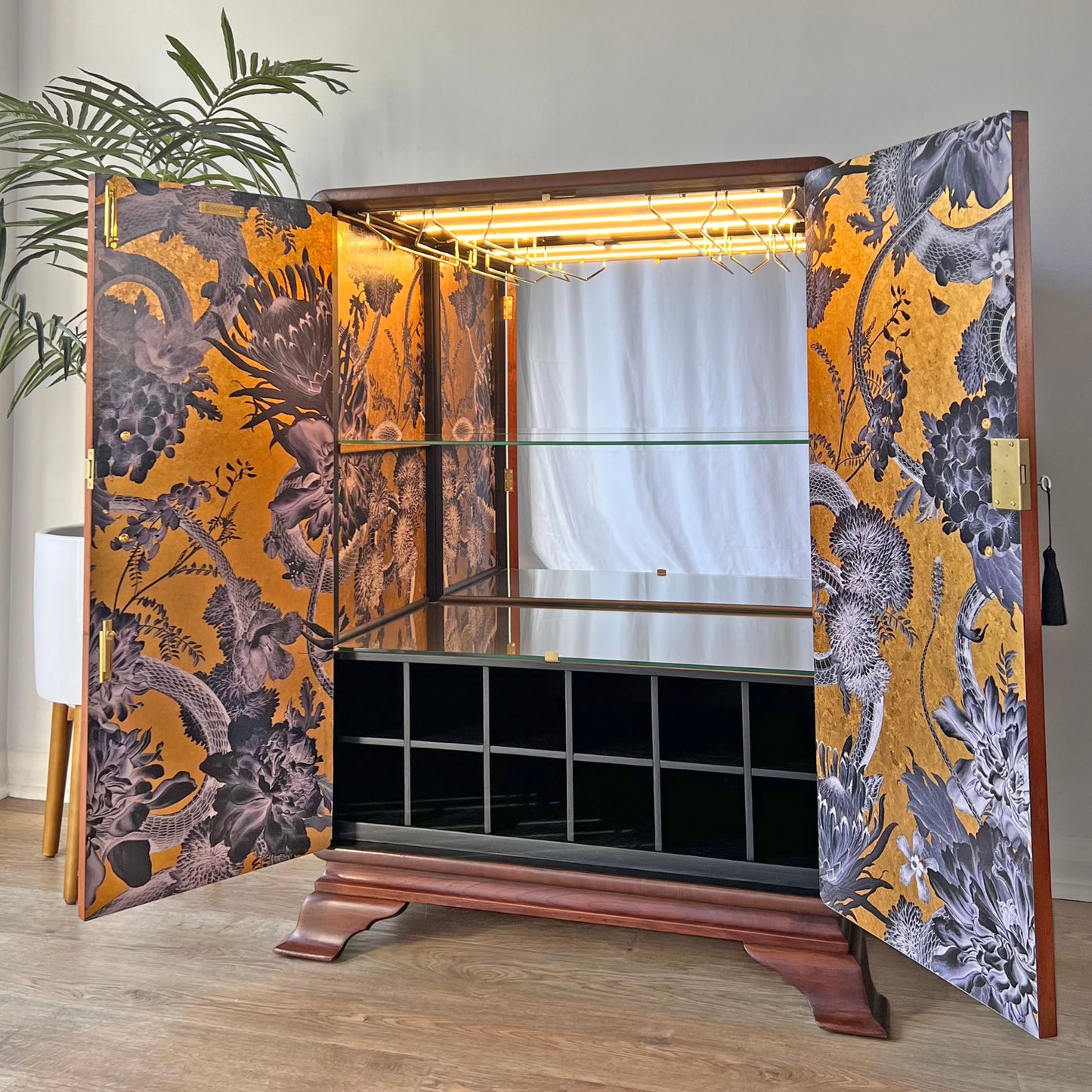 Open walnut drinks cabinet with empty glass shelves, integrated lighting, mirrored back panel and Divine Savages Forbidden Bloom wallpaper in real gold leaf.