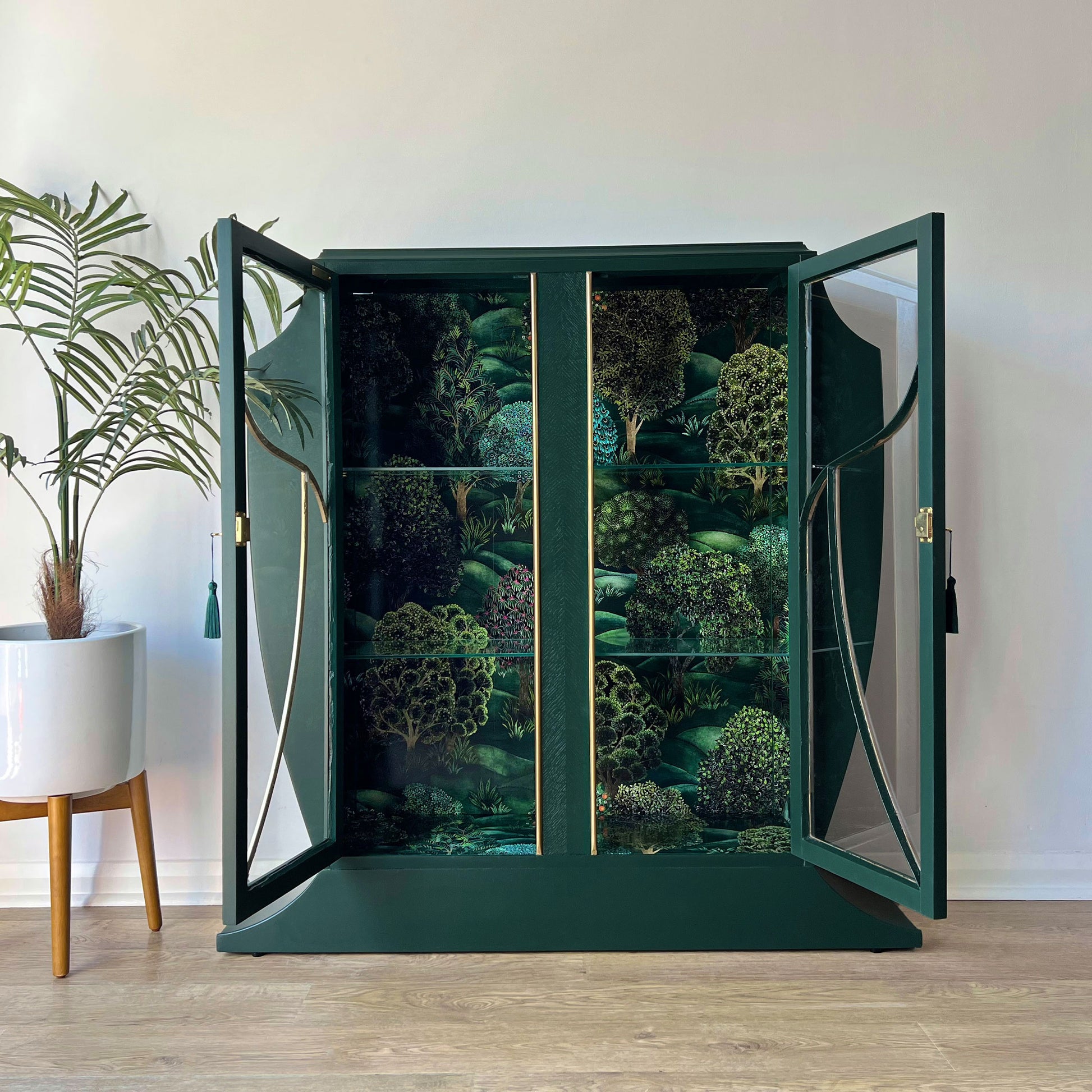Open green glazed drinks cabinet showing Cole & Son Forest wallpaper lining and glass shelving.