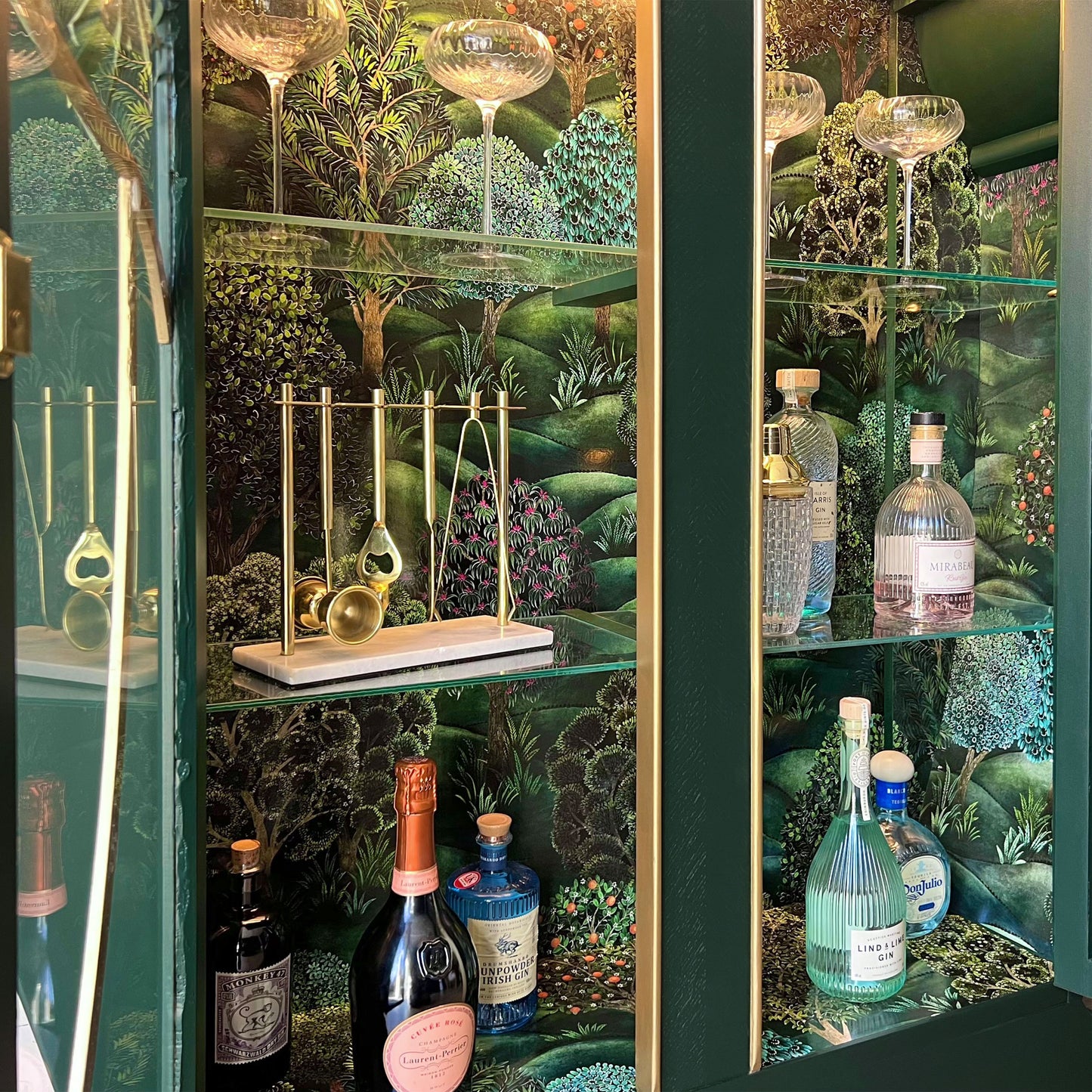 Detailed interior of green Art Deco bar cabinet with luxury bar tools and glassware set against Cole & Son Forest wallpaper.