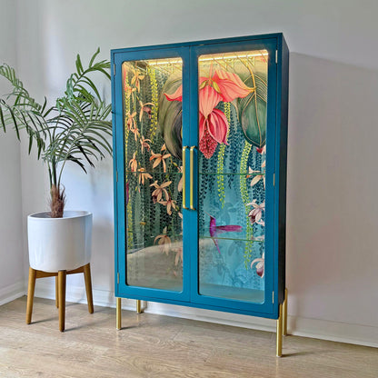 Angled image of a lit display cabinet with a botanical hummingbird wallpaper interior, teal frame, and gold handles.
