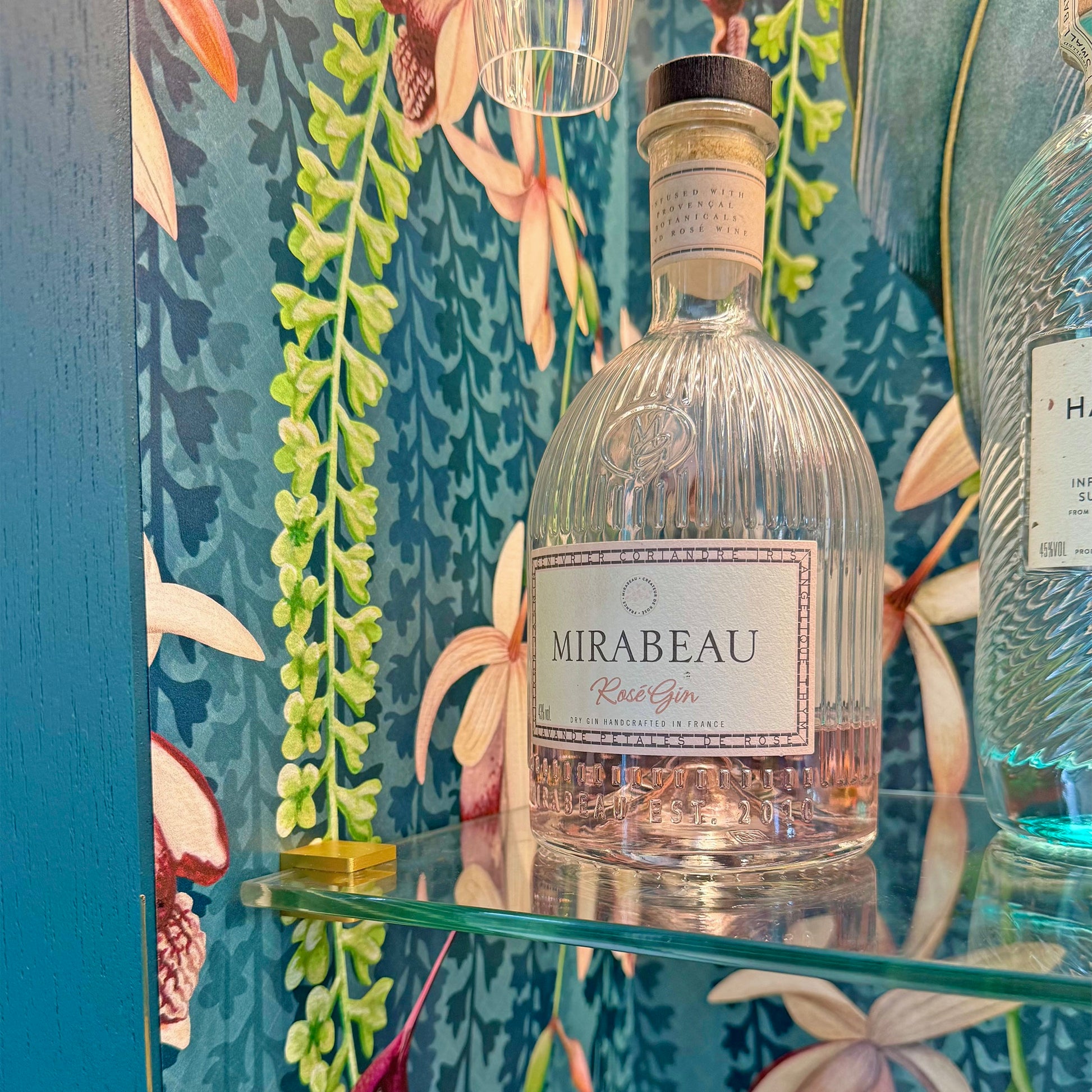 Detail shot of a Mirabeau Rose Gin bottle on a glass shelf with trailing orchid wallpaper as a backdrop.