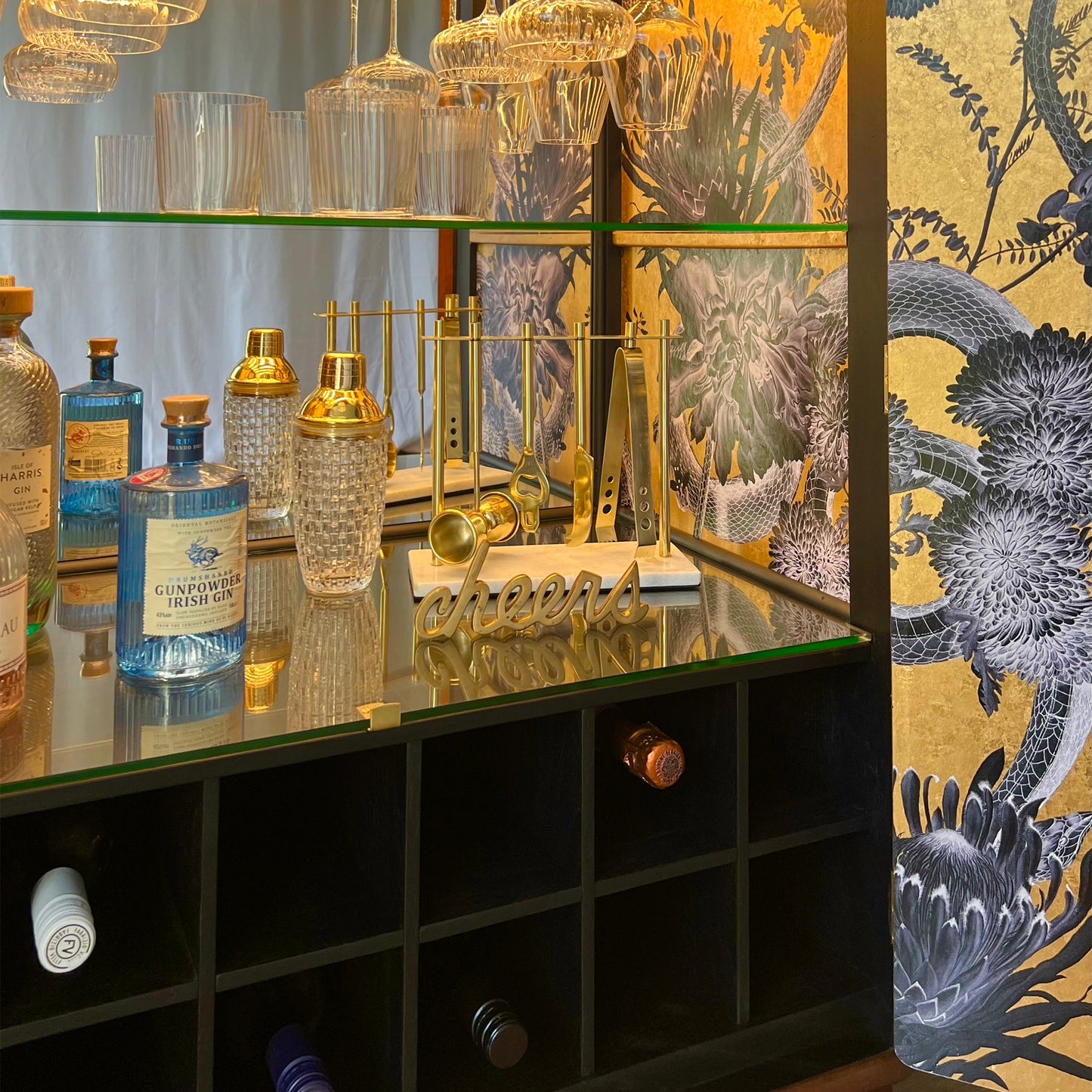 Curated barware and gin collection styled inside The Alchemist cabinet, with gold tools, crystal glassware, and 'cheers' sign on mirrored surface.