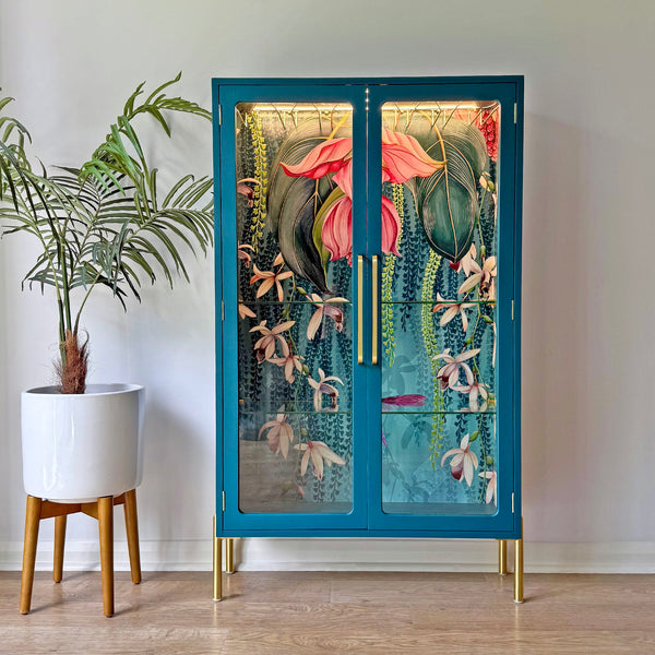 Front-facing image of a restored G Plan display cabinet painted in teal blue with gold legs, showcasing a botanical wallpaper interior featuring orchids and tropical foliage.